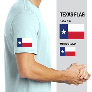 May include: A light blue t-shirt with a Texas flag patch on the sleeve. The flag is red, white, and blue with a single white star. The text "TEXAS FLAG" and "3.25 x 2 in" is displayed above a larger version of the flag. The text "MINI: 2 x 1.25 in" is displayed above a smaller version of the flag.