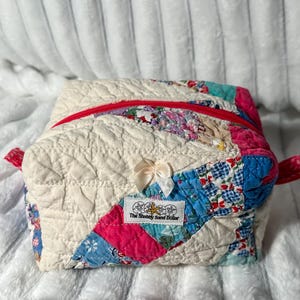 May include: A quilted fabric pouch featuring a patchwork design of cream, blue, and pink floral patterns. The bag has a red zipper closure, a small cream ribbon bow on the front, and a white rectangular label reading The Shabby Sand Dollar.