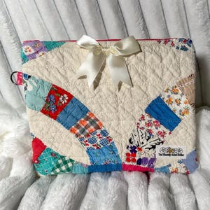 May include: A rectangular quilted pouch featuring a cream-colored center panel accented with a white satin ribbon bow. The pouch is bordered by a patchwork design of various floral and checkered fabric squares in red, blue, and green.