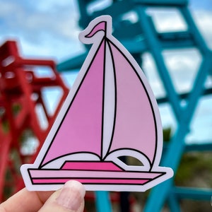 May include: A pink and white sailboat sticker with a black outline. The sticker is held in a hand against a blurred background of red and blue lifeguard stands.