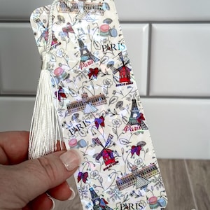 May include: A white bookmark with a tassel features a repeating pattern of Parisian landmarks, including the Eiffel Tower, a windmill, and the Louvre Museum. The bookmark is decorated with a floral design and the word "Paris" is repeated throughout the design.