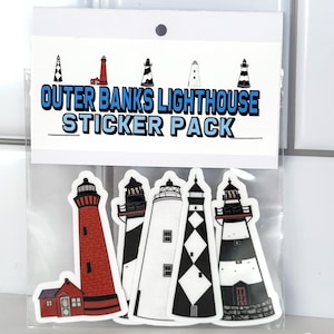 May include: A sticker pack featuring five Outer Banks lighthouse designs. The pack includes a red lighthouse, a white lighthouse with black stripes, a black and white lighthouse with a diamond pattern, a white lighthouse with black stripes, and a white lighthouse with black and red stripes. The sticker pack is packaged in a clear plastic bag with a white header card that reads "Outer Banks Lighthouse Sticker Pack".