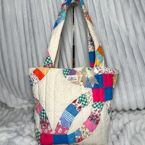May include: A quilted tote bag featuring a patchwork design with cream-colored fabric and various colorful floral and checkered patterns. The bag has two handles, a small white ribbon bow on the front, and a rectangular label reading The Sandy Sand Dollar.
