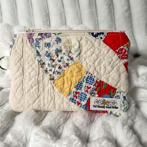 May include: A quilted zipper pouch featuring a cream-colored base with a diagonal patchwork design of floral, yellow, and red fabric patterns. A small white ribbon bow is attached to the front, and a label reads The Shabby Sand Dollar.
