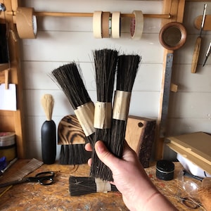 Op de afbeelding: Een verzameling donkerharige penselen, vastgebonden met bruin papier, wordt in een hand gehouden. De penselen zijn het centrale punt, met andere kunstbenodigdheden en gereedschappen zichtbaar op de achtergrond, waaronder rolletjes tape en een houten liniaal.