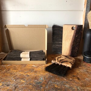 Op de afbeelding: Een set houten borstels met donkere haren, inclusief een rechthoekige borstel met een natuurlijke rand, een afgeronde borstel met een gebrande houten afwerking en een kartonnen doos met diverse bundels donkere haren samengebonden met papieren banden.