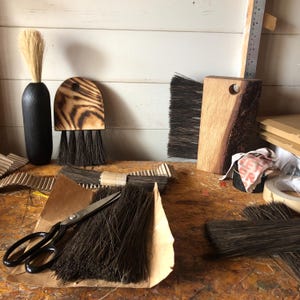 Op de afbeelding: Een verzameling handgemaakte borstels met donkere haren, waaronder een afgeronde houten borstel, een rechthoekige borstel met een natuurlijke rand en een borstel met een lange zwarte steel. Een metalen schaar ligt op een bundel losse donkere haren.