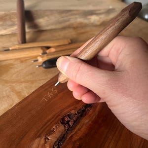 Può includere: Una penna di legno con punta sfaccettata è tenuta sopra una superficie di legno, con le lettere "TAS" incise. Sullo sfondo sono visibili altre penne di legno. La penna è di un ricco colore marrone.