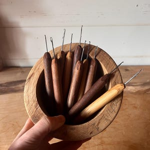 May include: A selection of wooden crochet hooks in a range of brown hues, presented in a round wooden bowl. The hooks have metal tips and are held in a hand. The bowl is crafted from light-coloured wood.