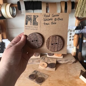Op de afbeelding: Twee handgesneden walnoten knopen met een gat van 4 mm worden gepresenteerd op een kartonnen achterkant. De knopen zijn rond met een textuur van houtnerf. De tekst op de kaart luidt "Hand Carved Walnut Buttons 4mm Hole" en het logo van de maker.