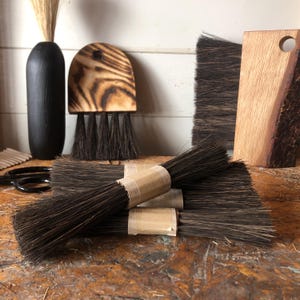 Op de afbeelding: Een verzameling donkerbruine borstels met stijve haren, waaronder verschillende bundels vastgebonden met bruin papieren tape, een halfronde borstel met een gebrand houten handvat en een rechthoekige borstel met een handvat van natuurlijk hout.