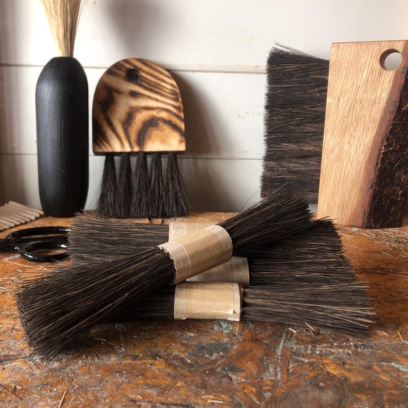 Op de afbeelding: Een verzameling donkerbruine borstels met stijve haren, waaronder verschillende bundels vastgebonden met bruin papieren tape, een halfronde borstel met een gebrand houten handvat en een rechthoekige borstel met een handvat van natuurlijk hout.