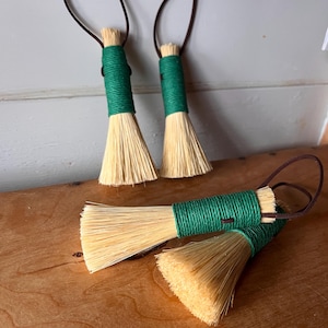 May include: Several small brushes with natural bristles, green twine-wrapped handles, and brown hanging loops. The brushes are displayed on a wooden surface. Each brush is approximately 15 cm in length.