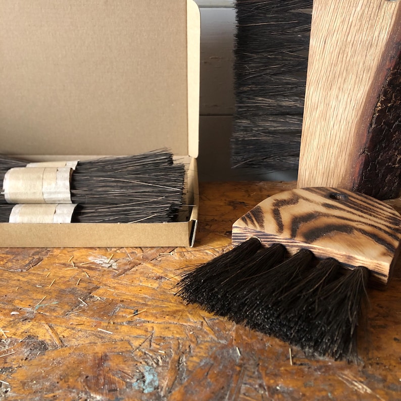 Op de afbeelding: Een set donkerbruine borstels met houten handvatten, sommige gebundeld met beige tape in een kartonnen doos en andere met geschroeide houtnerfpatronen, inclusief een gebogen borstelkop met een klein gaatje om op te hangen.