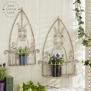 May include: Two white metal wall planters with a decorative arched design. Each planter has a shelf for displaying plants. The planters are hanging on a white wooden wall.
