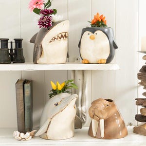 May include: Four ceramic animal planters, a shark, a penguin, a dolphin, and a walrus, are displayed on a white shelf. The planters are all white with brown, grey, and black accents. The shark planter has a flower arrangement with pink and purple flowers. The penguin planter has a small plant with orange flowers. The dolphin planter has a small plant with yellow flowers. The walrus planter is empty.