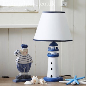 May include: A white and blue lighthouse-shaped table lamp with a white lampshade and a blue trim. The lamp is on a wooden surface with a wooden figurine of a sailor and seashells.