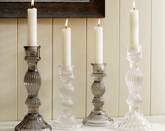 May include: Four glass candle holders with white candles. The candle holders are clear and have a textured design. Two of the candle holders are a darker gray color.