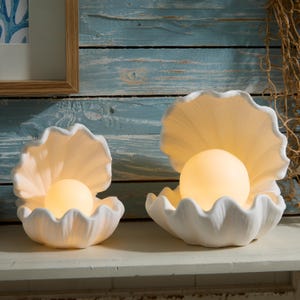 May include: Two white ceramic clam shell lamps of different sizes, each featuring a smooth, matte finish and an open shell design. Inside each shell sits a glowing, spherical light bulb that emits a warm, soft, and diffused yellow light.
