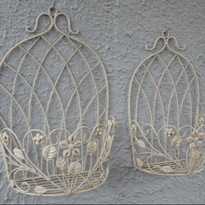 May include: Two off-white metal wall planters. The larger one is oval with a lattice design and floral details. The smaller one has a similar design. Both have a hanging loop at the top for display.