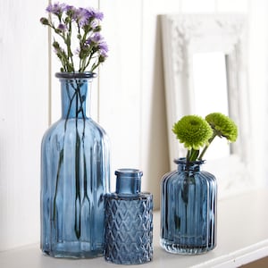 May include: Three blue glass vases of different sizes and designs. The tallest vase has a ribbed design and holds purple flowers. The middle vase has a diamond pattern and the smallest vase has a ribbed design and holds green flowers.