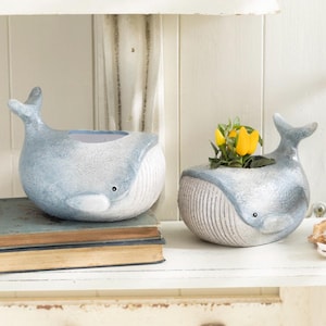 May include: Two blue and white whale-shaped planters, one with a small plant with yellow flowers, the other empty. The planters are sitting on a white shelf.