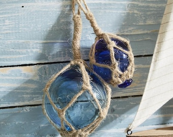 May include: Two blue glass floats wrapped in natural twine, hanging against a light blue wooden background. The floats are a popular nautical decor item.