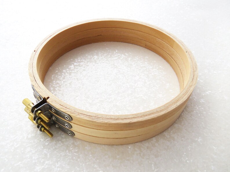 May include: A wooden embroidery hoop with a natural finish. The hoop is made of three layers of wood and has a metal tightening mechanism with three screws.