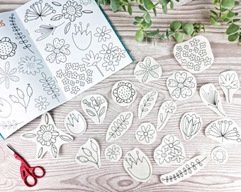 May include: A sketchbook with floral patterns and embroidered flower and leaf designs. A pair of red scissors is next to the sketchbook. The designs are black and white and are on a light wood surface.