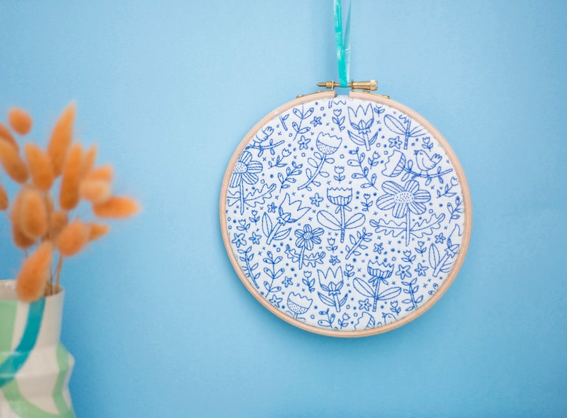 A floral doodle pattern in blue on white fabric, displayed in an embroidery hoop and hanging against a blue background with orange dried flowers in the foreground.