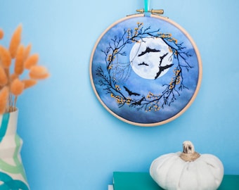 Puede incluir: Un aro de bordado azul y blanco con un diseño de luna y murciélagos. El aro está colgado en una pared azul claro. Una calabaza blanca se encuentra sobre una superficie verde en primer plano.