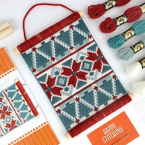 May include: A craft kit for creating a wall hanging with a geometric pattern of red, white, and blue snowflakes. The kit includes yarn, a needle, a wooden dowel, and a piece of Aida cloth. The kit also includes a small card that says "Happy Stitching!"