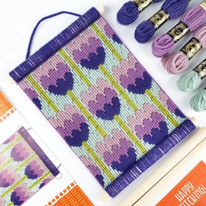 May include: A purple, green, and pink needlepoint wall hanging with a geometric pattern of hearts. The hanging is on a wooden dowel rod and comes with embroidery floss in various colors. The text "Happy Stitching!" is on a small orange card.