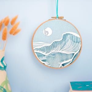 May include: An embroidered hoop art piece depicting ocean waves and a moon in shades of blue and white. The artwork is set within a wooden hoop and suspended by a teal ribbon. A decorative item for the home, measuring approximately 20 cm in diameter.