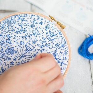 Hands working on a blue floral doodle embroidery design in a hoop with embroidery thread, needle, and scissors on a white wooden surface. Step-by-step instructions can be seen in the background.