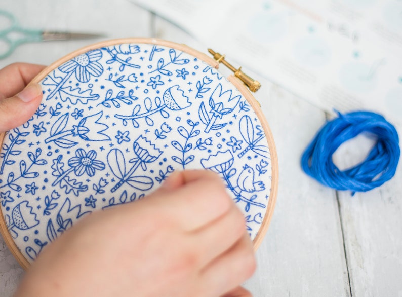 Hands working on a blue floral doodle embroidery design in a hoop with embroidery thread, needle, and scissors on a white wooden surface. Step-by-step instructions can be seen in the background.