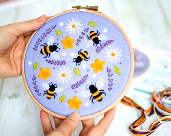 May include: A purple embroidery hoop with a floral design featuring bees, flowers, and leaves. The embroidery is done in shades of yellow, white, and purple.