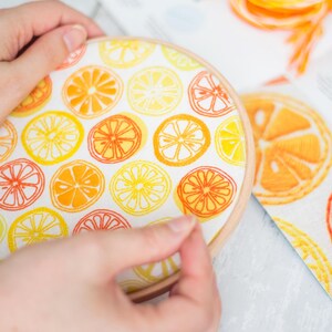 May include: A close-up of a hand holding a round embroidery hoop with a white fabric inside. The fabric is printed with a pattern of yellow and orange citrus slices.