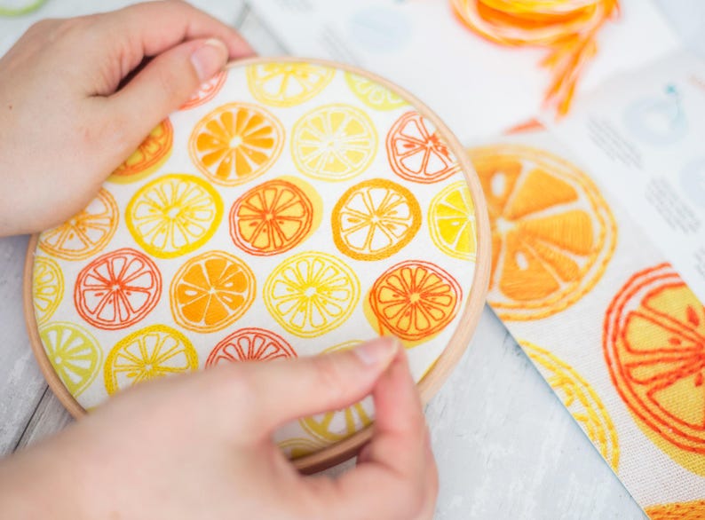 May include: A close-up of a hand holding a round embroidery hoop with a white fabric inside. The fabric is printed with a pattern of yellow and orange citrus slices.