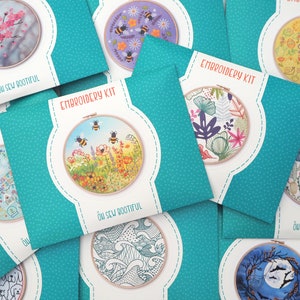 Assorted embroidery kits laid out on a surface. Each kit features a different design, including bees, flowers, cacti, waves, and more, with packaging showing the finished embroidery in a hoop. The kits are in teal packaging with white polka dots.
