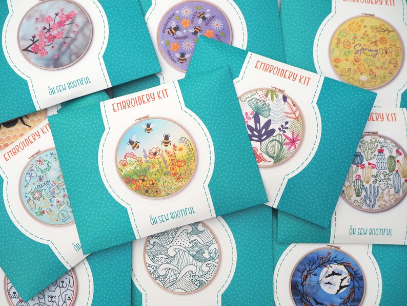 Assorted embroidery kits laid out on a surface. Each kit features a different design, including bees, flowers, cacti, waves, and more, with packaging showing the finished embroidery in a hoop. The kits are in teal packaging with white polka dots.