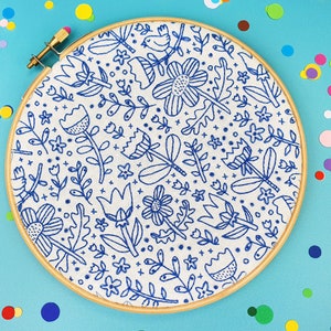 An embroidery hoop displaying a blue and white floral doodle pattern on fabric, set against a turquoise background scattered with colourful confetti dots. This playful design features various flowers, leaves, and small decorative elements.