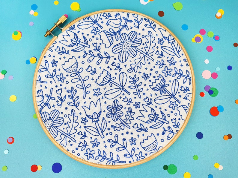 An embroidery hoop displaying a blue and white floral doodle pattern on fabric, set against a turquoise background scattered with colourful confetti dots. This playful design features various flowers, leaves, and small decorative elements.