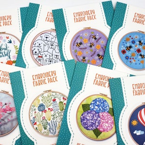 May include: Eight embroidery hoop kits with pre-printed fabric designs. The designs include a floral wreath with the text "be kind to yourself", a group of dogs, bees and flowers, bees and flowers on a blue background, a cherry blossom tree, a cactus garden, a cluster of hydrangeas, and a hot air balloon with mountains.