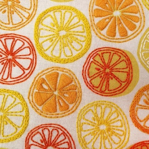 May include: Close-up of a white fabric with embroidered yellow and orange citrus fruit designs. The fabric is textured and has a subtle sheen.