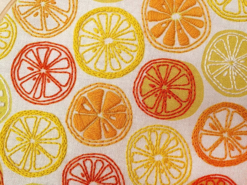 May include: Close-up of a white fabric with embroidered yellow and orange citrus fruit designs. The fabric is textured and has a subtle sheen.