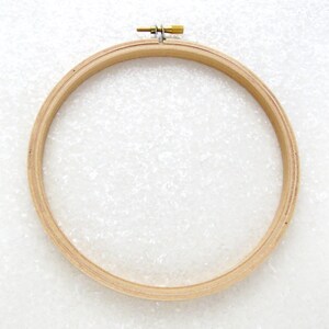 May include: A wooden embroidery hoop with a brass screw tightening mechanism. The hoop is round and has a natural wood finish.