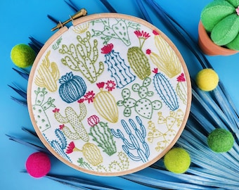 May include: A white embroidery hoop with a colorful cactus pattern. The hoop is surrounded by green, blue, and pink pom-poms.