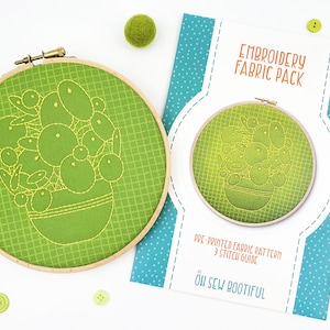 May include: A green embroidery hoop with a pre-printed design of a potted plant with yellow stitching. The hoop is on a white surface with green buttons and balls of yarn. The packaging for the embroidery kit is in the background and says "Embroidery Fabric Pack" and "Oh Sew Bootiful".