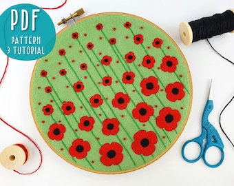 May include: A green embroidery hoop with a design of red poppies and green stems. The hoop is on a white surface with red and black embroidery floss and a pair of blue embroidery scissors.
